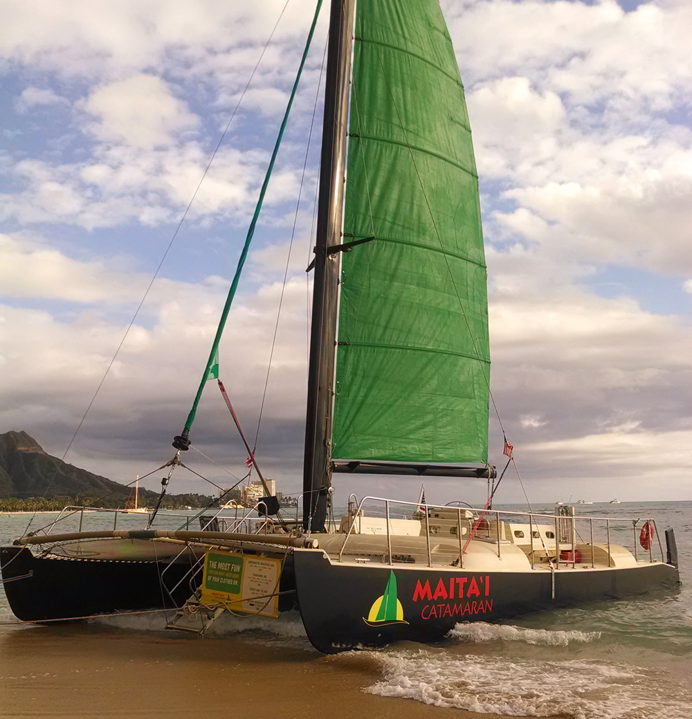 The authentic Maitaʻi Catamaran on Waikiki Beach, with its distinctive green sail and dark hulls